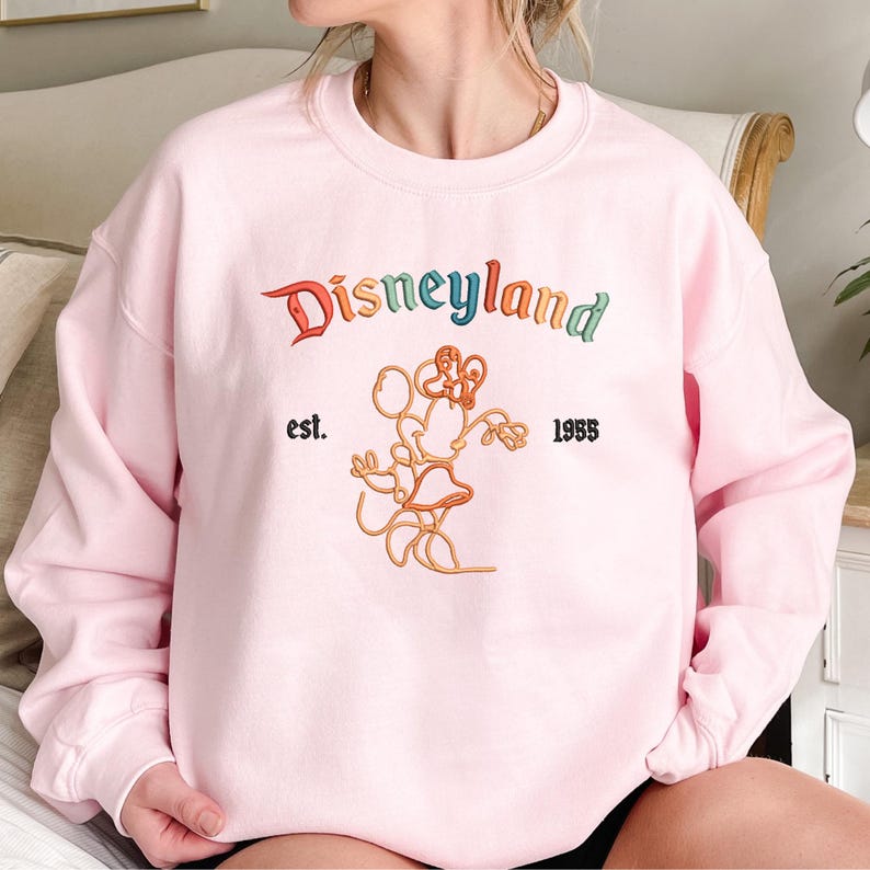 Może przedstawiać: Jasnor&oacute;żowa bluza z okrągłym dekoltem i wyhaftowanym logo Disneyland w wielokolorowych literach. Pod napisem znajduje się pomarańczowy kontur Myszki Minnie oraz czarny wyhaftowany napis est. 1955 po obu stronach.