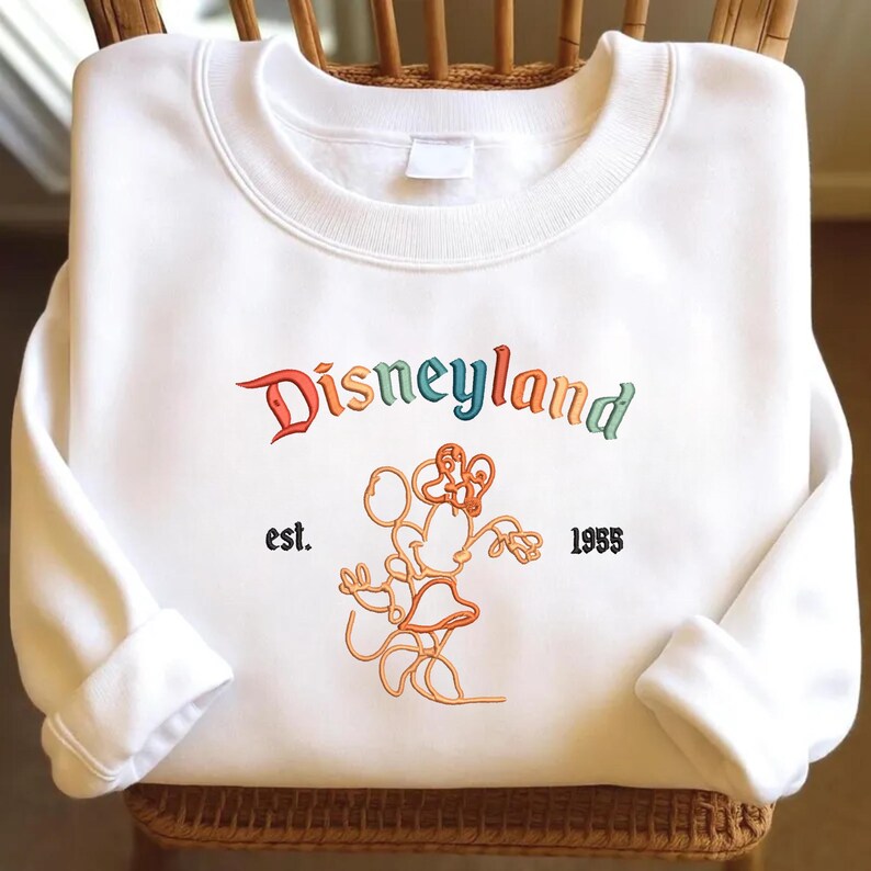 Może przedstawiać: Biała bluza z okrągłym dekoltem z wyhaftowanym wielokolorowym logo Disneyland w zakrzywionej czcionce. Poniżej tekstu znajduje się pomarańczowa ilustracja liniowa myszy, otoczona czarnym wyhaftowanym napisem est. oraz 1955.