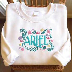 Op de afbeelding: Een wit sweatshirt met lange mouwen en een geborduurd ontwerp op de borst. De tekst ARIEL is gestikt in talingblauw, omringd door kleurrijke geborduurde motieven waaronder roze zeesterren, gele sterren, paarse schelpen en talingblauwe zeewierranken.