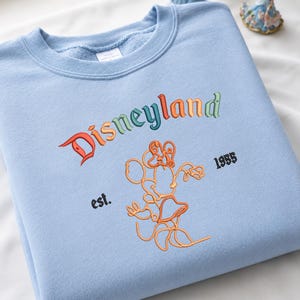 Może przedstawiać: Jasnoniebieska bluza z okrągłym dekoltem z wyhaftowanym słowem Disneyland wielokolorowymi literami. Pod napisem znajduje się pomarańczowy haft przedstawiający zarys myszy z kresk&oacute;wki oraz czarny haft z napisem est. 1955 po bokach.