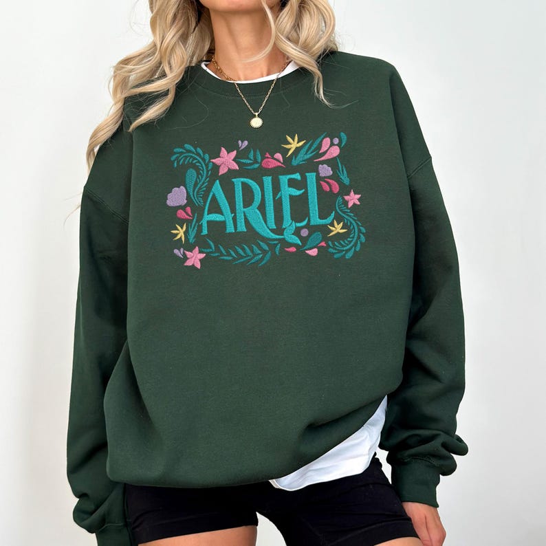 Op de afbeelding: Een donker bosgroen sweatshirt met ronde hals en de naam ARIEL in groenblauwe letters, omringd door een geborduurd ontwerp van roze bloemen, gele sterren en groenblauwe bladranken. Het kledingstuk wordt gedragen over een wit shirt en zwarte short.