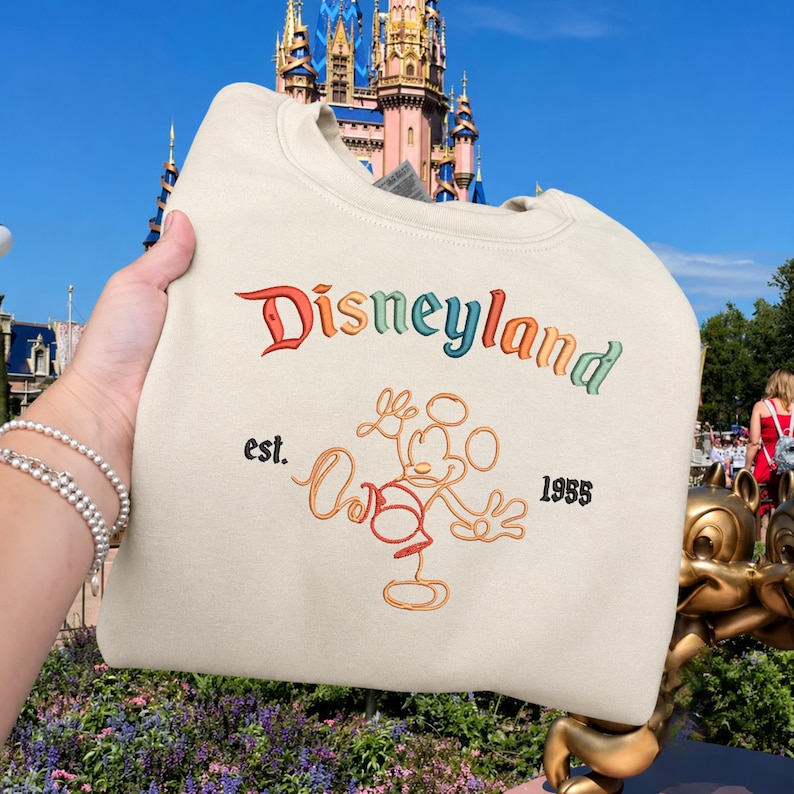 May include: A cream-coloured crewneck sweatshirt featuring the word Disneyland embroidered in a multi-coloured gradient font. Below the text is an orange outline of Mickey Mouse with the words est. and 1955 embroidered in black on either side.