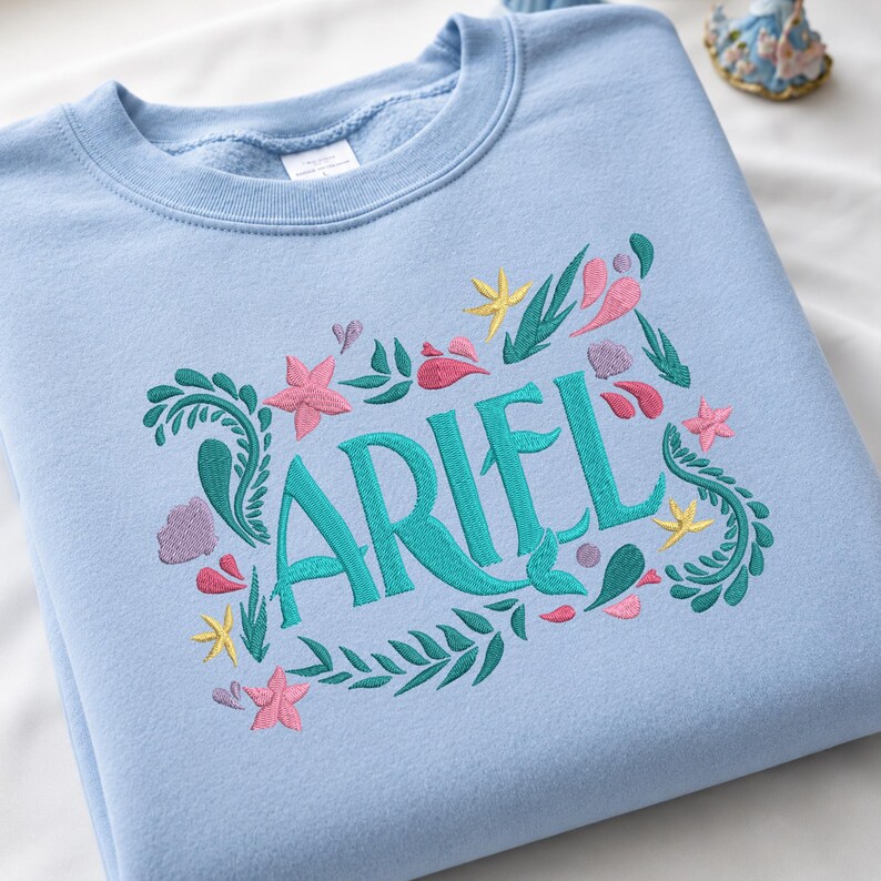 Op de afbeelding: Een lichtblauw sweatshirt met ronde hals en een geborduurd ontwerp met de naam ARIEL in grote groenblauwe letters. De tekst is omringd door kleurrijke geborduurde motieven, waaronder roze zeesterren, gele sterren, paarse schelpen en groen zeewier.