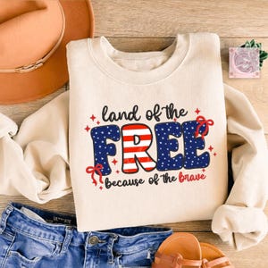 May include: Cream-colored sweatshirt with the text "land of the FREE because of the brave" in red, white, and blue. The word "FREE" has a blue star pattern and red stripes. A pair of blue jeans and a brown hat are also visible.
