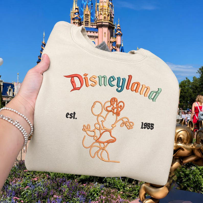Może przedstawiać: Kremowa bluza z okrągłym dekoltem z napisem Disneyland wyhaftowanym wielokolorową czcionką gradientową. Pod tekstem znajduje się pomarańczowa ilustracja liniowa Myszki Minnie, otoczona czarnym wyhaftowanym napisem est. oraz 1955.