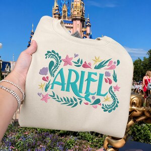 Op de afbeelding: Een cr&egrave;mekleurig sweatshirt met ronde hals en de naam ARIEL geborduurd in groenblauw. Het ontwerp is omringd door geborduurde botanische en aquatische elementen in tinten groenblauw, roze en geel, waaronder bladeren, zeesterren en schelpen.