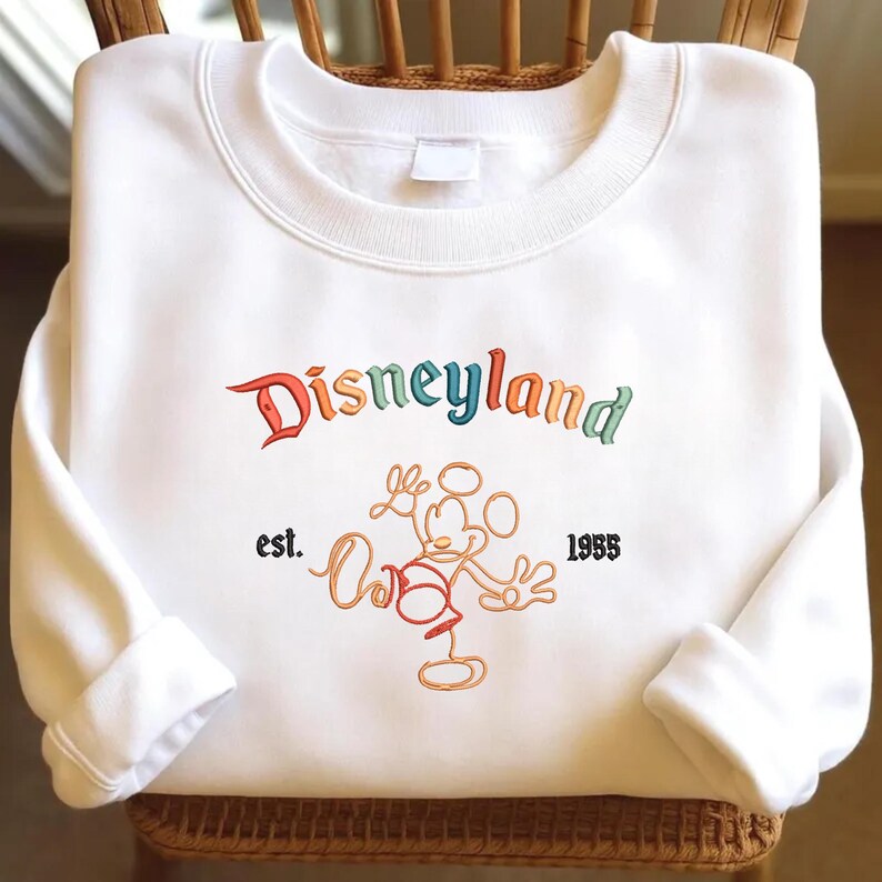 May include: A white crewneck sweatshirt featuring the word Disneyland embroidered in multicoloured letters. Below the text is an orange outline of a cartoon mouse character with the text est. 1955 embroidered in black on either side of the figure.