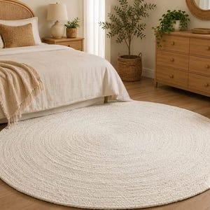 May include: A large, circular, off-white area rug featuring a thick, braided, or coiled texture. The rug is positioned on a wooden floor in front of a bed, showcasing a consistent, woven pattern that radiates from the center to the outer edge.