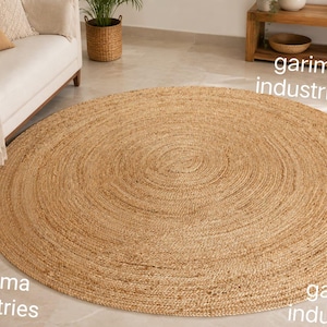 May include: A large, round, natural-colored woven jute rug featuring a concentric circular pattern. The rug has a textured, braided surface and is displayed in a room setting. The text "garima industries" is visible in three locations on the image.
