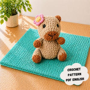 Puede incluir: Un oso de peluche tejido a ganchillo con una flor rosa y amarilla en la cabeza está sentado sobre un cuadrado turquesa texturizado. El oso es de color canela con detalles marrones. El texto en forma de nube dice "CROCHET PATTERN PDF ENGLISH".
