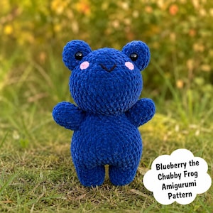 May include: A handmade, blue amigurumi frog with black eyes and pink cheeks is positioned on grass. The frog has a round body and small arms and legs. Text on a white cloud-shaped graphic reads "Blueberry the Chubby Frog Amigurumi Pattern."