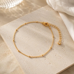 May include: A gold-toned chain bracelet featuring evenly spaced small metal beads along the length. The bracelet includes a lobster claw clasp, an adjustable extender chain, and a single small clear round stone charm at the end of the extender.