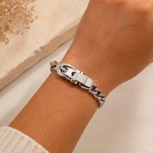 May include: A silver-toned metal chain bracelet featuring a central buckle-style clasp mechanism. The bracelet consists of interlocking polished links that form a continuous band, secured by a functional metal buckle closure in the centre.