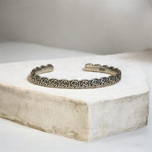 May include: A silver cuff bracelet with a repeating rose pattern. The open bracelet is displayed on a white surface. The roses are detailed, adding an elegant touch to the jewellery.