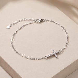 May include: A silver-toned chain bracelet featuring a small cross charm encrusted with clear, sparkling stones. The bracelet includes a delicate link chain, a secure lobster claw clasp, and an adjustable extender chain with a small flat metal tag.