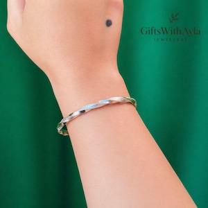 May include: A silver bangle with a twisted pattern. The bracelet is displayed on a wrist, set against a green backdrop. The jewellery is from GiftsWithAyla.