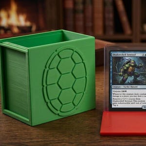 May include: Three-piece card game storage set. A green box with a turtle shell design, a red card display, and a red box with a dice tray. The set holds cards and dice. The card display shows a card with the text "Shadowshell Sentinel".