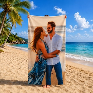 May include: A rectangular beach towel featuring a printed photograph of two people embracing on a sandy shore. The image shows a person with long hair in a blue patterned dress and a person with a beard in a white shirt and blue pants.