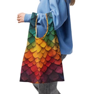 May include: A reusable tote bag featuring a vibrant, colorful pattern resembling overlapping scales. The design transitions from green to yellow, orange, red, and purple. The bag is held by a person wearing a blue hoodie.
