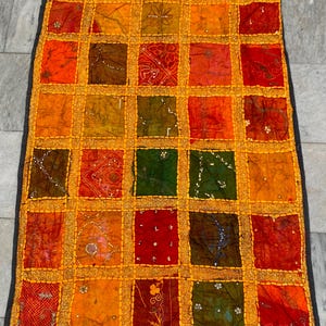 May include: A rectangular patchwork textile with a grid of colorful squares in shades of orange, red, green, and brown. Each square has a unique pattern and is bordered by gold trim. The edges are finished with a dark gray border.