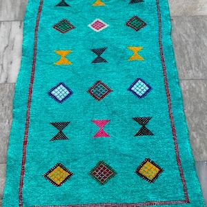 Puede incluir: Una alfombra rectangular turquesa con borde rojo y flecos. Presenta un patrón repetido de diamantes y formas de reloj de arena en amarillo, rosa, negro y verde. La alfombra es probablemente hecha a mano.