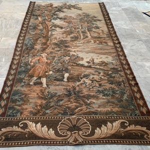 May include: A rectangular tapestry featuring a hunting scene with figures in period clothing, dogs, and a forest backdrop. The tapestry has a brown, beige, and green color palette with a decorative border and a detailed design.