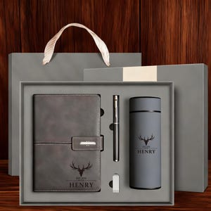 May include: A grey gift set containing a notebook with a deer graphic and the text "EST. 2026 HENRY", a matching pen, a small metal clip, and a cylindrical grey tumbler featuring the same deer graphic and the name "HENRY" printed on the front.
