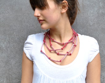 May include: A woman wearing a white shirt with a layered pink and red necklace. The necklace is made of multiple strands of knotted cord.