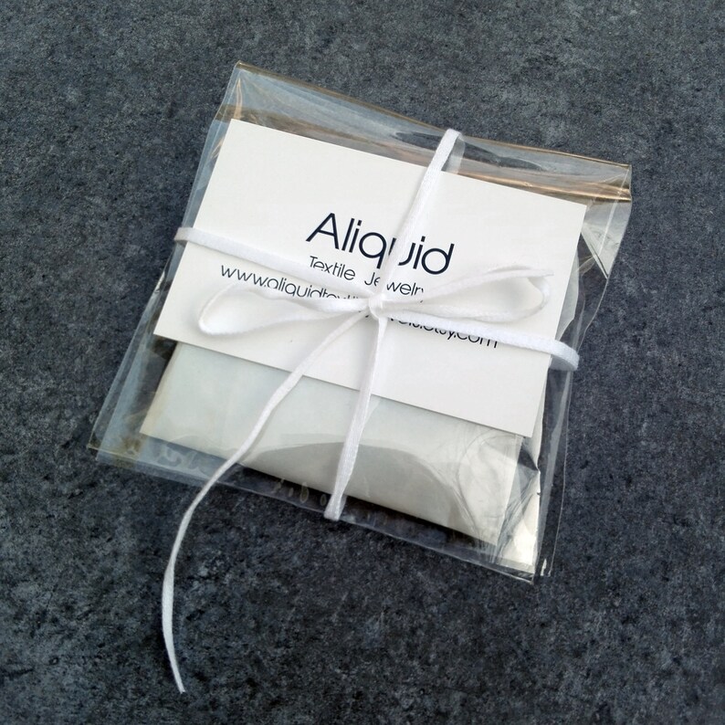 Peut inclure: Une enveloppe blanche avec un ruban blanc nou&eacute; autour. L'enveloppe contient une carte blanche avec le texte "Aliquid Textile Jewelry www.aliquidjewelry.com" imprim&eacute; dessus.