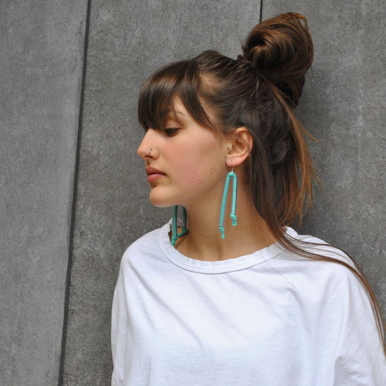 Peut inclure: Une femme avec de longs cheveux bruns portant un t-shirt blanc et des boucles d'oreilles pendantes turquoise. Ses cheveux sont tir&eacute;s en arri&egrave;re en un chignon.
