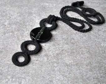May include: A black crocheted necklace with a large black button pendant. The necklace is made of multiple loops of crocheted cord.