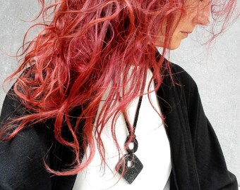 May include: A woman with long, curly, red hair is wearing a black cardigan and a white shirt. She is wearing a black pendant necklace with a geometric shape.