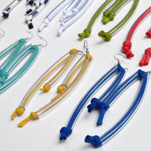Peut inclure: Une paire de boucles d'oreilles color&eacute;es en plastique transparent et en tissu. Les boucles d'oreilles sont longues et pendantes, avec des n&oelig;uds aux extr&eacute;mit&eacute;s du tissu. Les couleurs incluent le rouge, l'orange, le jaune, le vert, le bleu et le blanc.