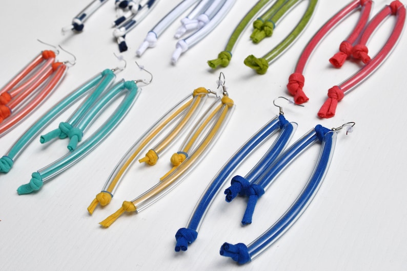 Peut inclure: Une paire de boucles d'oreilles color&eacute;es en plastique transparent et en tissu. Les boucles d'oreilles sont longues et pendantes, avec des n&oelig;uds aux extr&eacute;mit&eacute;s du tissu. Les couleurs incluent le rouge, l'orange, le jaune, le vert, le bleu et le blanc.