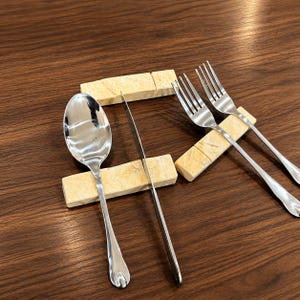 May include: A stainless steel spoon, knife, and forks rest on rectangular beige stone holders. The silverware is arranged on a dark brown wooden surface, creating a contrast in textures and colors. The utensils are clean and reflect the light.