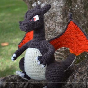 Könnte beinhalten: Handgehäkelter Drache in Dunkelgrau mit orangefarbenen und roten Flügeln. Der Drache hat einen weißen Bauch und weiße Akzente an den Füßen. Die Augen sind weiß mit roten Pupillen. Der Drache sitzt auf einem Ast.