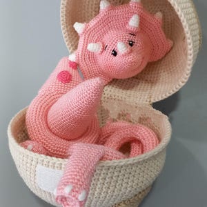 Puede incluir: Un peluche de dinosaurio triceratops rosa hecho a mano con detalles blancos, metido en una cesta de ganchillo. El dinosaurio tiene una expresión amigable y está hecho de hilo suave. La cesta es de color crema.