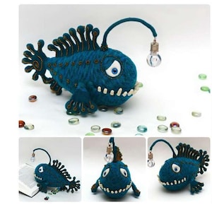 May include: A handmade, teal-colored anglerfish plush toy with a light bulb lure. The plush has white teeth, a detailed fin, and a whimsical design. The toy is surrounded by decorative glass beads.