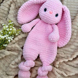 Könnte beinhalten: Ein handgefertigtes rosa Häkel-Hasen-Plüschtier mit langen Ohren, blauen Augen und einer rosa Nase. Der Hase hat ein weiches, strukturiertes Aussehen und geknotete Füße. Das Spielzeug ist als kuscheliger Begleiter konzipiert.