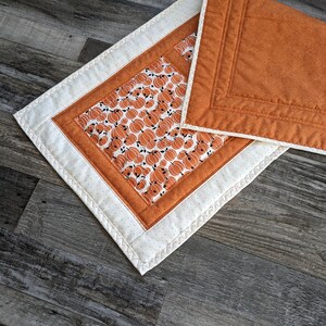 May include: A white and orange quilted table runner with a pumpkin print. The runner has a white border with a stitched design and a white backing with orange stitching.