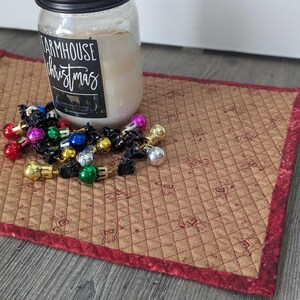 Puede incluir: Una vela en tarro de cristal transparente con tapa negra y una etiqueta que dice "Farmhouse Christmas". La vela está sobre un mantel individual acolchado con borde rojo. Pequeños adornos navideños de colores están esparcidos alrededor de la vela.