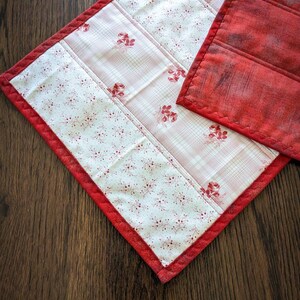 May include: Red and white quilted table runner with a floral pattern. The runner has a red border and is made of fabric.