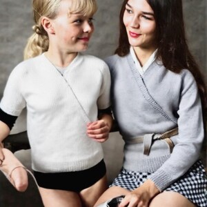 May include: Two people are seated, wearing knit wrap sweaters. One wears a white sweater with black shorts, the other a gray sweater and a black and white checkered skirt. A ballet slipper is held.