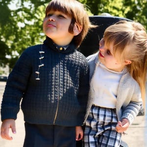 May include: Two children are shown in a daytime outdoor setting. One child wears a dark blue textured knit sweater with a gold zipper and dark pants. The other child wears a light gray cardigan, a white shirt, and black and white plaid pants.