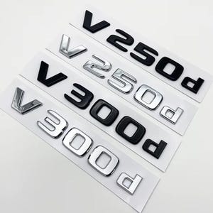 Könnte beinhalten: Vier Auto-Emblem-Aufkleber in einer Reihe auf weißem Trägerpapier. Das erste und dritte Emblem sind mattschwarz mit der Aufschrift V250d und V300d. Das zweite und vierte Emblem sind verchromt mit der Aufschrift V250d und V300d.