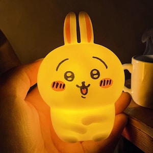 Puede incluir: Una luz de noche amarilla con forma de conejito con una cara sonriente, mejillas rosadas y orejas erguidas. La luz está encendida y el conejito se sostiene en una mano. El fondo es oscuro, con una taza blanca visible.