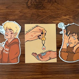 May include: Three stickers on a wooden surface. One sticker features a blonde person in a brown hoodie smoking a cigarette. Another sticker shows a key being handed to a bandaged hand. The third sticker depicts a brown-haired person in a brown hoodie with a cigarette and the text "I'm fine."