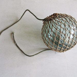 Op de afbeelding: Een vintage glazen visdrijver, omhuld door een geweven net, met een stuk touw eraan bevestigd. De heldere glazen bol heeft een lichte blauwgroene tint. Het net is lichtbruin van kleur. Het touw is een gedempt groen.
