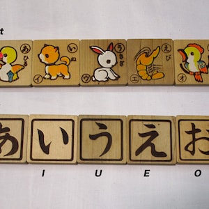 Puede incluir: Cinco bloques de madera con caracteres japoneses e ilustraciones de un pato, un gato, un conejo, un camarón y un pájaro. La parte posterior de los bloques muestra los caracteres japoneses para 'a', 'i', 'u', 'e' y 'o'.