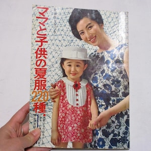 Könnte beinhalten: Vintage japanisches Zeitschriften-Cover mit einer Frau und einem kleinen Mädchen. Das Mädchen trägt ein rotes Blumenkleid und einen weißen Hut. Die Frau trägt ein blaues Blumenkleid. Der Zeitschriften-Titel ist auf Japanisch.